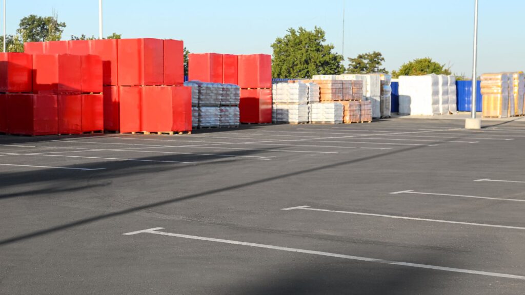 An empty parking lot with several stacks of red and white pallets and other materials neatly arranged in the background hints at a bustling Contractor's Asphalt warehouse nearby.